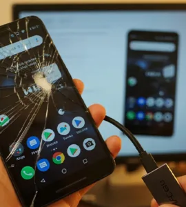 Téléphone avec écran cassé : image ou photo d'un smartphone Android avec un écran fortement fissuré au premier plan. L’écran affiche une interface légèrement visible (comme des icônes ou un écran d’accueil flou). Une main tient une souris branchée au téléphone avec un câble OTG.