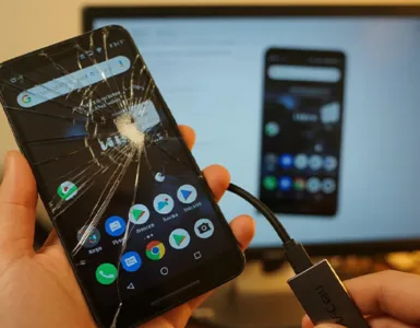 Téléphone avec écran cassé : image ou photo d'un smartphone Android avec un écran fortement fissuré au premier plan. L’écran affiche une interface légèrement visible (comme des icônes ou un écran d’accueil flou). Une main tient une souris branchée au téléphone avec un câble OTG.