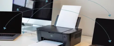 Partage d'une imprimante : image ou photo d'un bureau moderne avec deux ordinateurs portables et un ordinateur de bureau connectés à une même imprimante Wi-Fi au centre. Des lignes de connexion discrètes illustrent le réseau local entre les appareils et l'imprimante.